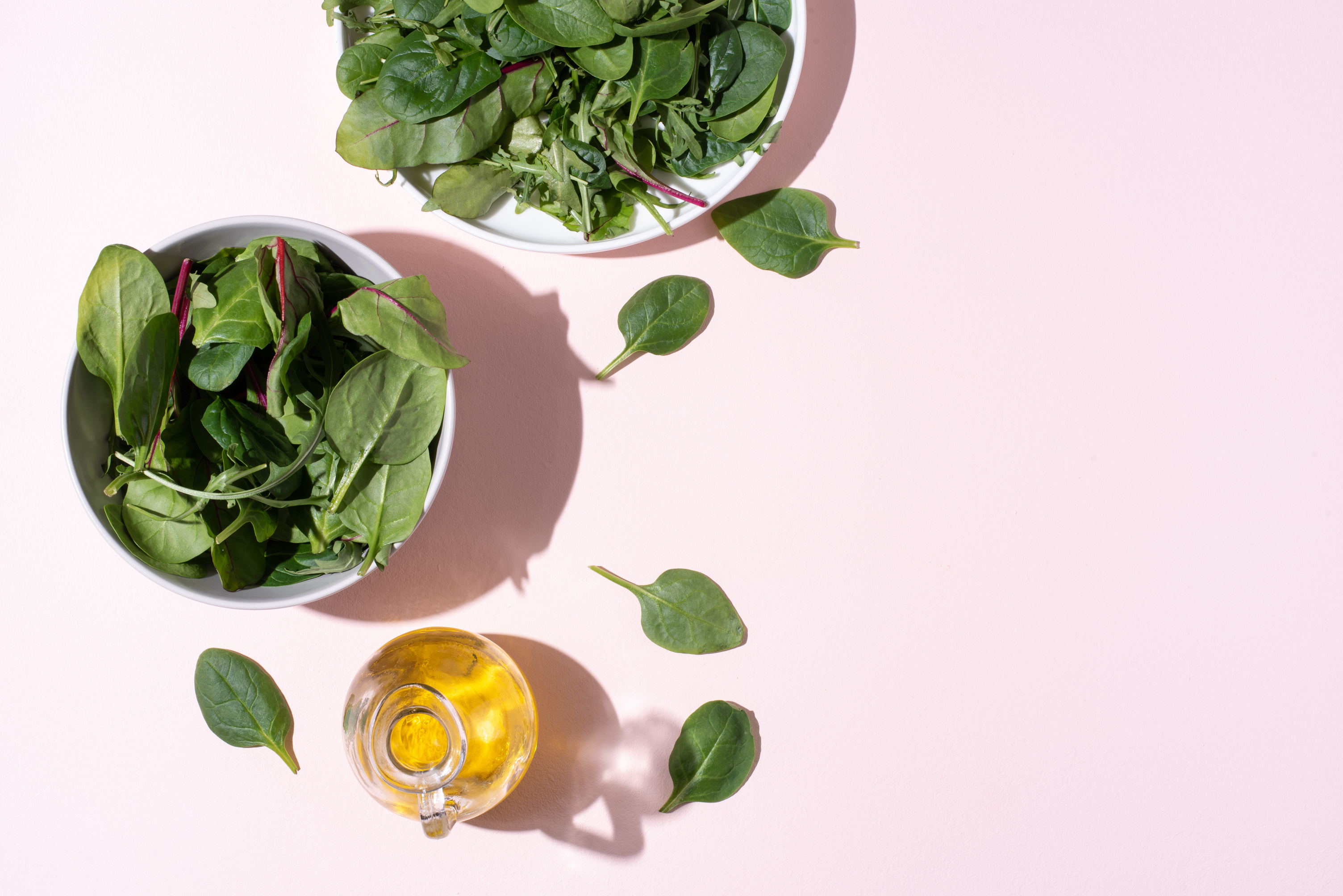 Plates with leafy greens, olive oil on pink background in sunlight, vegetarian food, healthy eating.